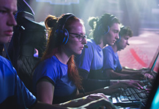 Concentrated young female esportsman gamer in glasses playing shooter on computer while sitting amidst teammates during gaming tournament